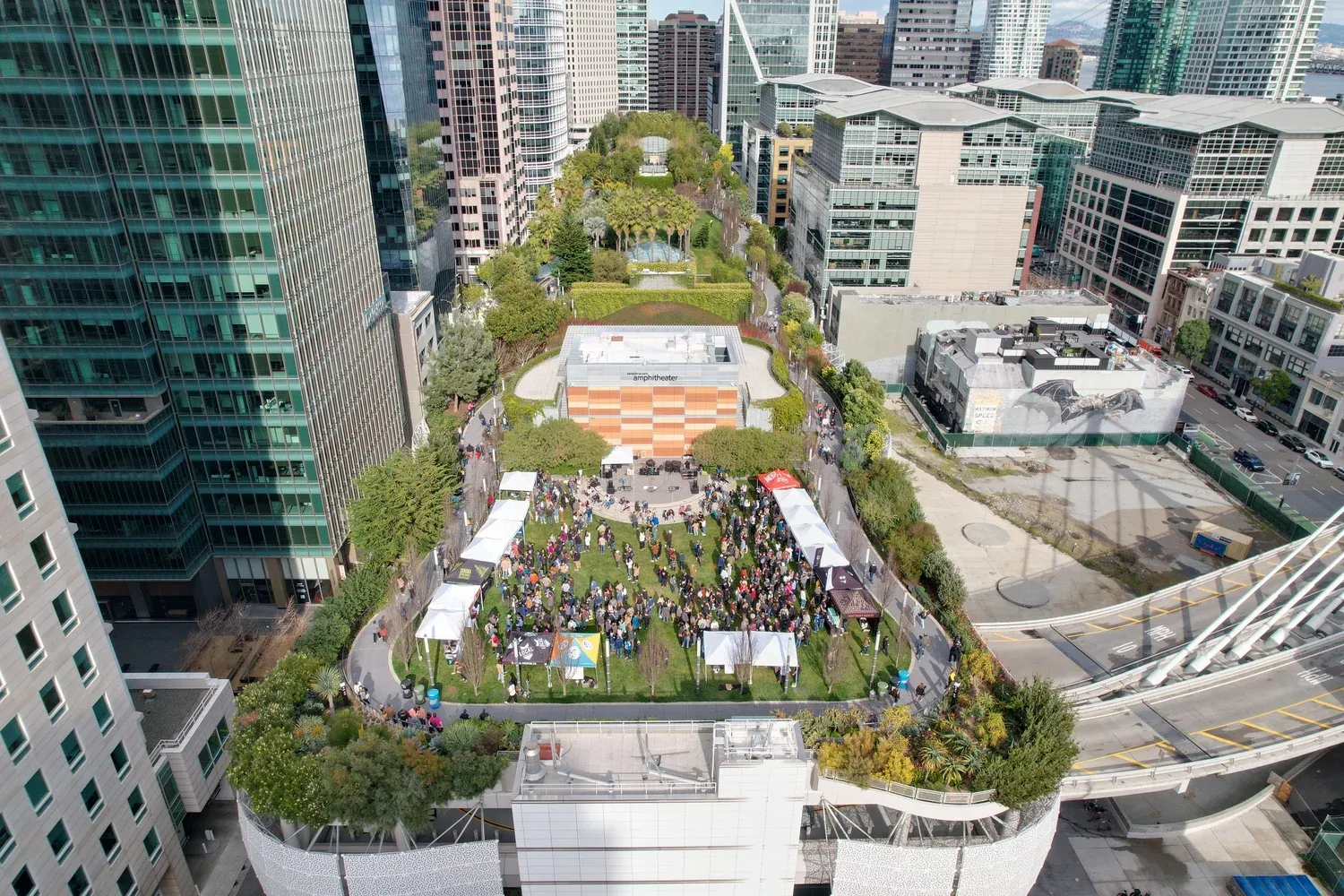 Aerial view of the Salesforce Park beer garden with white tents, trees, and crowds, surrounded by downtown San Francisco towers.